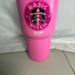 May include: A bright pink insulated tumbler with a silver rim. The front features a circular logo with a black silhouette of a witch wearing a hat, surrounded by the text BASIC WITCH. Below the logo, the text Sandy Cheeks is printed in white.