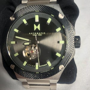 May include: A stainless steel automatic watch with a black dial and luminous hands and markers. The watch features a visible movement and a textured bezel. The watch band is also stainless steel.