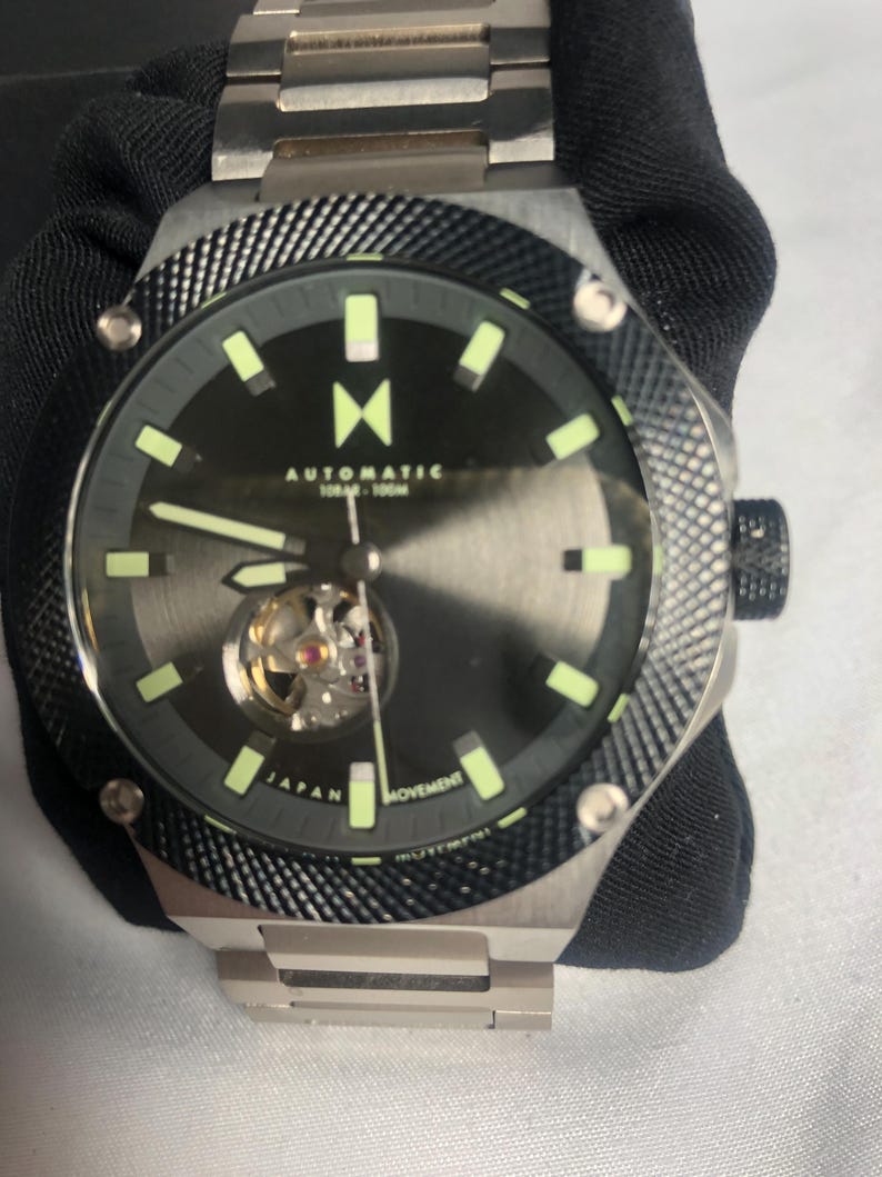 May include: A stainless steel automatic watch with a black dial and luminous hands and markers. The watch features a visible movement and a textured bezel. The watch band is also stainless steel.