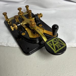 May include: A telegraph key featuring a black rectangular base with brass components and a black knob. The knob displays a yellow triangle logo containing the letters SAC and the number 70. The device includes multiple adjustable brass screws.
