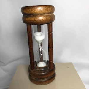 May include: A wooden hourglass with a clear glass center. The hourglass has a dark brown wooden frame with a rounded top and bottom. White sand is visible inside the glass, and the hourglass is sitting on a beige surface.