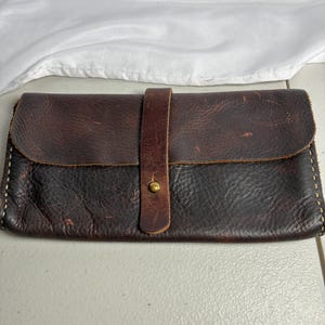 May include: A rectangular dark brown leather wallet featuring a textured surface, a fold-over flap, and a vertical leather strap secured with a single brass-colored metal snap button. The edges are finished with visible light-colored stitching.
