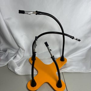 May include: A yellow X-shaped base with three flexible black gooseneck arms attached. Each arm features a silver metal alligator clip at the end with black rubber tips. The base is flat and provides a stable foundation for the adjustable arms.