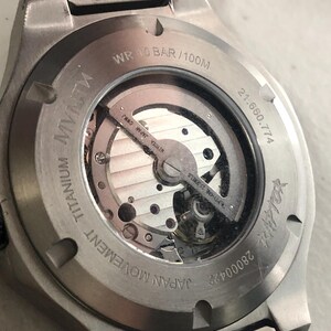 May include: A close-up of the rear of a titanium wristwatch. The watch has a transparent window revealing the Japanese movement. Text includes "JAPAN MOVEMENT TITANIUM", "WR 10 BAR/100M", and "MIYOTA JAPAN CWMJ". The watch has a silver metal strap.