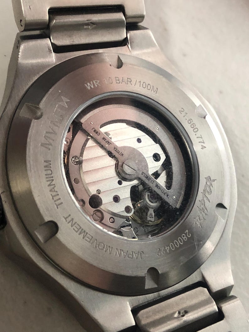 May include: A close-up of the rear of a titanium wristwatch. The watch has a transparent window revealing the Japanese movement. Text includes "JAPAN MOVEMENT TITANIUM", "WR 10 BAR/100M", and "MIYOTA JAPAN CWMJ". The watch has a silver metal strap.