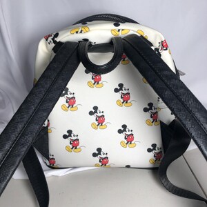 K&ouml;nnte beinhalten: Wei&szlig;er Rucksack mit einem sich wiederholenden Muster von Mickey Mouse in roten Shorts, gelben Schuhen und schwarzen Handschuhen. Der Rucksack hat schwarze Tr&auml;ger und einen schwarzen oberen Griff. Der Rei&szlig;verschluss ist schwarz.