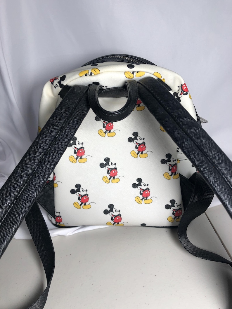 K&ouml;nnte beinhalten: Wei&szlig;er Rucksack mit einem sich wiederholenden Muster von Mickey Mouse in roten Shorts, gelben Schuhen und schwarzen Handschuhen. Der Rucksack hat schwarze Tr&auml;ger und einen schwarzen oberen Griff. Der Rei&szlig;verschluss ist schwarz.