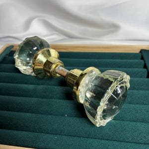 May include: A vintage-style door knob set featuring two clear, faceted glass knobs connected by a central metal spindle. The glass knobs have a multi-sided, reflective surface, and the connecting hardware is finished in a polished gold-tone metal.
