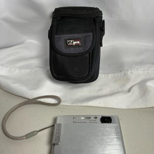 May include: A silver digital camera with a 5x optical zoom lens and Carl Zeiss Vario-Tessar branding, featuring a grey wrist strap. Accompanying the camera is a black Vidpro protective carrying case with a textured fabric exterior and a front flap.