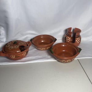 May include: A set of four terracotta serving dishes. The set includes a lidded bowl with a spoon, two open bowls, and a napkin holder. Each piece is decorated with white floral accents and green trim.