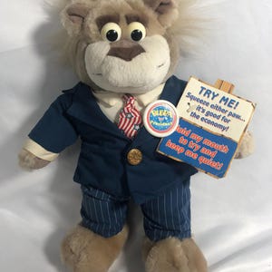 May include: A plush lion toy wearing a navy blue suit jacket, a white collared shirt, a red and white striped tie, and pinstriped trousers. A round button reads Bubba for President. A sign attached to the hand reads TRY ME! Squeeze either paw... it's good for the economy! Hold my mouth to try and keep me quiet!