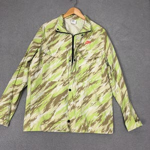 May include: A long-sleeved jacket with a green, white, and brown camouflage pattern. The jacket has a zipper closure, black buttons, and a small red logo on the chest. It is hanging on a wooden hanger.