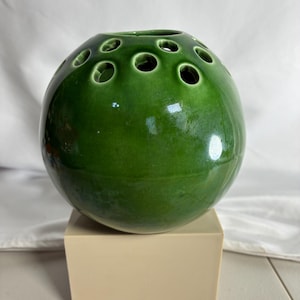 May include: A spherical, glossy green ceramic flower frog vase featuring a series of circular openings across the top surface for arranging stems, resting on a solid, light beige rectangular base.