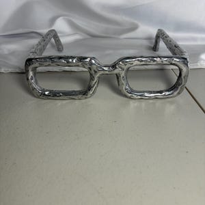 May include: A pair of silver-toned eyeglasses featuring a thick, textured frame with a rugged, hammered surface finish. The rectangular lenses are absent, leaving the metallic rims open. The temples are curved at the ends for a secure fit.