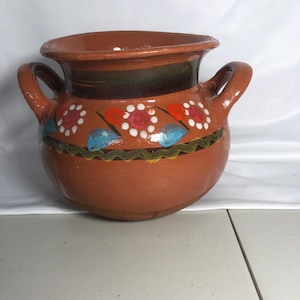 May include: A terracotta-colored ceramic pot with two handles. The pot features a decorative band with red, white, and blue floral designs. The rim and handles are a darker shade of brown.