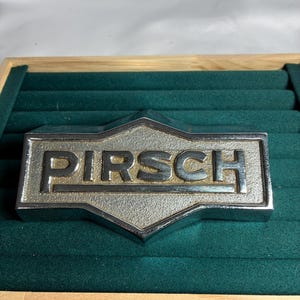 May include: A rectangular, metallic silver-toned emblem featuring the word PIRSCH in raised, bold capital letters. The background of the text area has a textured, granular finish, while the border and letters are smooth and reflective.