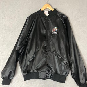 May include: Black satin bomber jacket with a button-up front, two front pockets, and a ribbed collar, cuffs, and hem. The jacket features a colorful embroidered logo on the chest. The jacket is hanging on a wooden hanger.