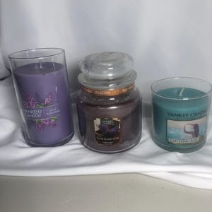 May include: Three Yankee Candles in glass containers. One is a tall, cylindrical candle in a lilac color with "Lilac Blossoms" text. The middle candle is a jar candle with a lid, and the label reads "Dried Lavender & Oak." The third candle is teal with "Catching Rays" text.