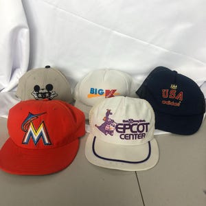May include: A collection of baseball caps in various colors. One is red with a white and blue logo, another is white with a purple and white graphic, and a navy blue cap with "USA" text. Other caps include a gray Mickey Mouse cap.