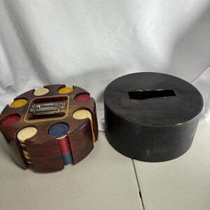 May include: A circular wooden poker chip carousel with a metal handle, containing stacks of red, blue, yellow, and white chips. Beside it sits a separate, solid black circular lid with a rectangular slot in the center for storage.