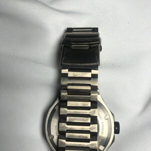 May include: A silver-toned MVMT watch with a titanium band. The watch face is visible, with the text "MVMT" on the clasp. The band is made of rectangular links. The watch has a black dial and a black crown.