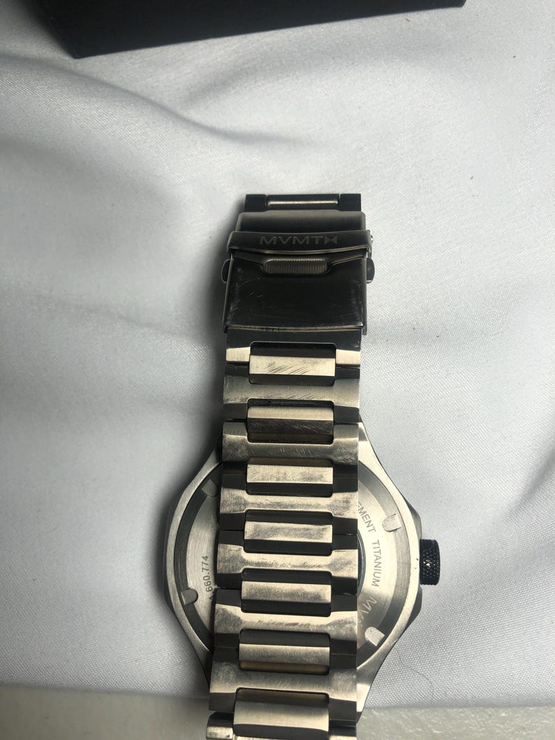 May include: A silver-toned MVMT watch with a titanium band. The watch face is visible, with the text "MVMT" on the clasp. The band is made of rectangular links. The watch has a black dial and a black crown.