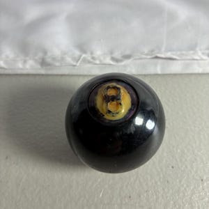 May include: A spherical black billiard ball featuring a circular inset on the top surface. The inset contains a yellow background with a dark, weathered number eight printed in the center. The surface of the ball is smooth and reflective.