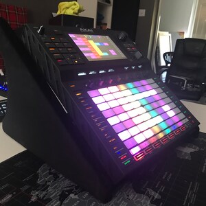 以下が含まれることがあります： カラフルなバックライト付きパッドグリッドと、音楽シーケンスを表示する画面を備えた、黒いAkai Force音楽制作ワークステーション。デバイスは黒いスタンドに角度を付けて置かれ、黒と白の世界地図のマウスパッドの上に置かれています。