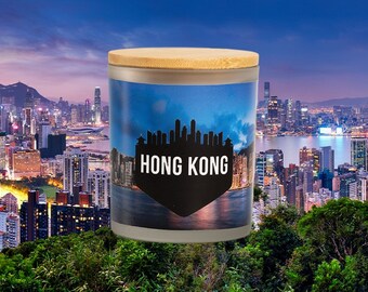 May include: A candle with a bamboo lid and a cityscape of Hong Kong printed on the side. The candle is in front of a blurry background of the Hong Kong skyline.