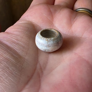 May include: A miniature, round ceramic vessel with a wide, open top. The exterior features a mottled, textured finish in shades of off-white, cream, and light brown, with a smooth, rounded base and a slightly darker, earthy rim around the opening.
