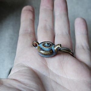 May include: A miniature teapot ring with a blue and brown glazed ceramic finish. The ring features a small, detailed teapot design with a handle and spout. The ring is set on a silver band.