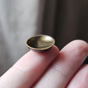 May include: A miniature brass bowl with a dark interior and a raised rim. The bowl has a round shape and a warm, golden colour. It appears to be handmade. The bowl is small enough to fit on a fingertip.