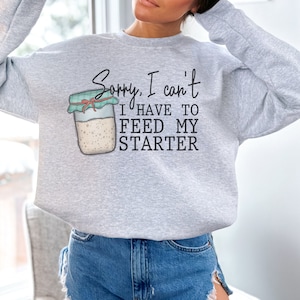 May include: A light gray sweatshirt with the text "Sorry, I can't I have to feed my starter" and an illustration of a jar of sourdough starter. The sweatshirt is a crew neck style.