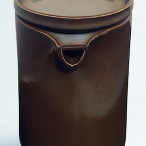 May include: A cylindrical dark brown ceramic pitcher with a smooth, glossy finish. The vessel features a flat, circular lid that sits flush with the top rim and a small, integrated spout located just below the lid, creating a seamless profile.