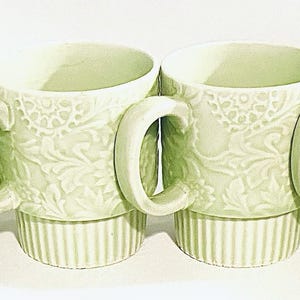May include: Four light green ceramic mugs with a floral embossed design. Each mug has a curved handle and a ribbed base. The mugs are stacked and arranged in a row, perfect for serving coffee or tea.