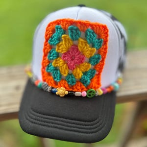 May include: A white and black trucker hat featuring a square, multi-colored crochet granny square patch on the front panel. The base of the front panel is adorned with a string of assorted colorful beads, including a small turtle and peace sign.