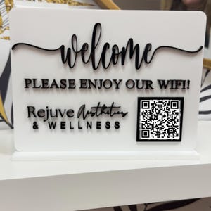 May include: White sign with black text that reads "Welcome, Please Enjoy Our Wifi! Rejuve Aesthetics & Wellness". A black and white QR code is in the bottom right corner.