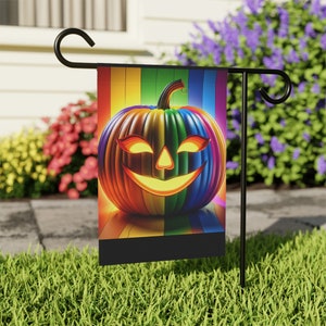 Może przedstawiać: Kolorowa flaga ogrodowa na Halloween z tęczową dynią z świecącą twarzą Jack-o'-lantern. Flaga jest eksponowana na czarnym metalowym stojaku w ogrodzie z kwiatami w tle. Projekt dyni jest żywy i przyciąga wzrok.