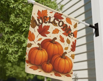 May include: A beige outdoor flag with the word "WELCOME" in brown script, surrounded by orange and brown autumn leaves and pumpkins. The flag is hanging on a white wall next to green foliage.