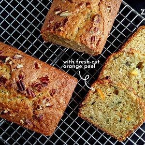 May include: Three loaves of zucchini pecan bread with fresh-cut orange peel on a wire rack. The bread is golden brown and has a rustic appearance. "Zucchini Pecan" is written in white text on a black background.