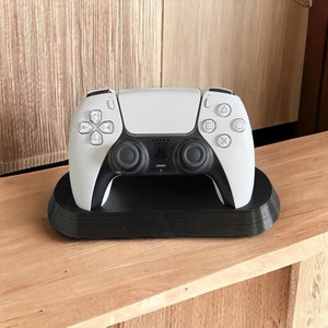 May include: A white PlayStation 5 DualSense wireless controller is sitting on a black charging stand. The controller has a black PlayStation logo and black buttons.
