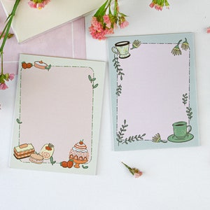 Pode incluir: Dois blocos de notas retangulares com fundos rosa pastel e azul claro. Um bloco de notas apresenta ilustrações de sobremesas, o outro tem um design de xícara de chá. Ambos têm bordas de folhas e flores verdes. Flores rosas e artigos de papelaria no fundo.