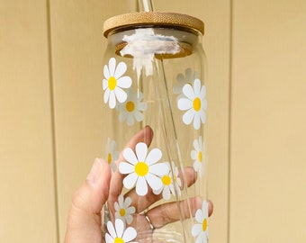May include: A clear glass reusable water bottle with a bamboo lid and a glass straw. The bottle is decorated with white daisies with yellow centers.