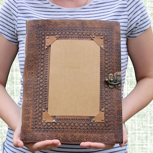 May include: A brown leather journal with intricate embossed floral and geometric patterns. The journal features a rectangular, blank, tan-colored paper insert, brass corner accents, and a decorative clasp. The cover is a rich, dark brown color.