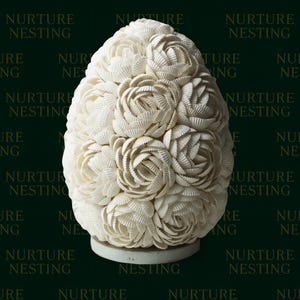 Puede incluir: Una escultura decorativa en forma de huevo, compuesta por numerosas conchas blancas, dispuestas para parecer rosas. La escultura se asienta sobre una pequeña base redonda blanca. Las palabras "NURTURE NESTING" están impresas en el fondo verde oscuro.