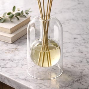 Könnte beinhalten: Ein klarer Glasdiffusor mit rundem Boden und gewölbtem Deckel, gefüllt mit einer hellgelben Flüssigkeit und Holzstäbchen. Der Diffusor steht auf einer Marmoroberfläche, im Hintergrund ein Stapel Bücher und Grün.