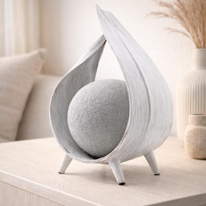 May include: A white wooden sculpture featuring a teardrop design, holding a grey sphere. The sculpture has three small legs and a distressed finish. The sphere is made of woven threads. The sculpture is a decorative item.