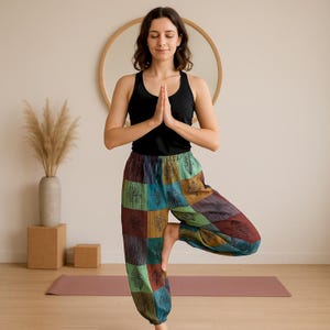 May include: A person in a black vest top and patchwork yoga trousers in a tree pose. The trousers have a colourful patchwork design with teal, brown, and green squares. The background includes a mirror, a vase with dried plants, and a yoga mat.