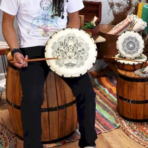 Op de afbeelding: Een set handgemaakte drums met een cr&egrave;mekleurig drumvel met een zwart Hamsa-handontwerp. De drums staan op houten vaten. Een drumstok ligt op het drumvel. De drums zijn geschikt voor meditatie en muziek.