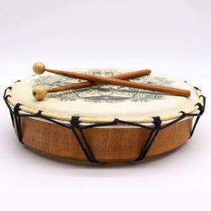 Op de afbeelding: Een kleine, ronde trommel met een lichtgekleurd vel met een zwart ontwerp. De trommel heeft een houten frame en is bespannen met zwart touw. Twee houten drumstokken met afgeronde uiteinden liggen erop.